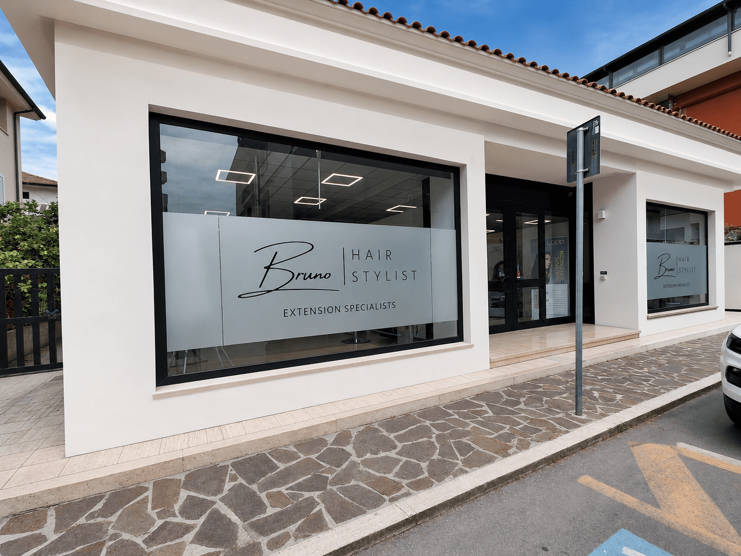 Facciata bianca del salone Bruno Hair Stylist con ampie vetrate e insegne per extension.